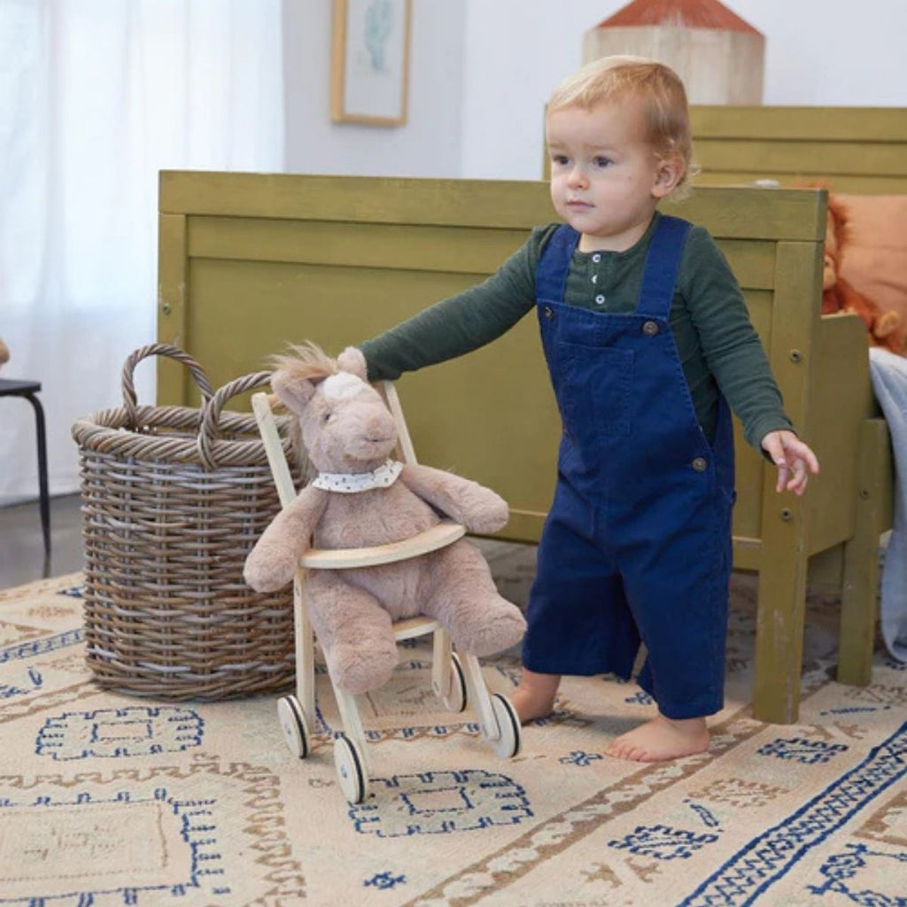 Child in blue overalls standing next to a plush toy on a wooden stool in a room with a rug and furniture.