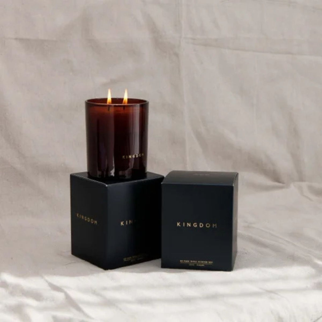Candle in a dark glass container with a black box labeled 'Kingdom' on a light gray background