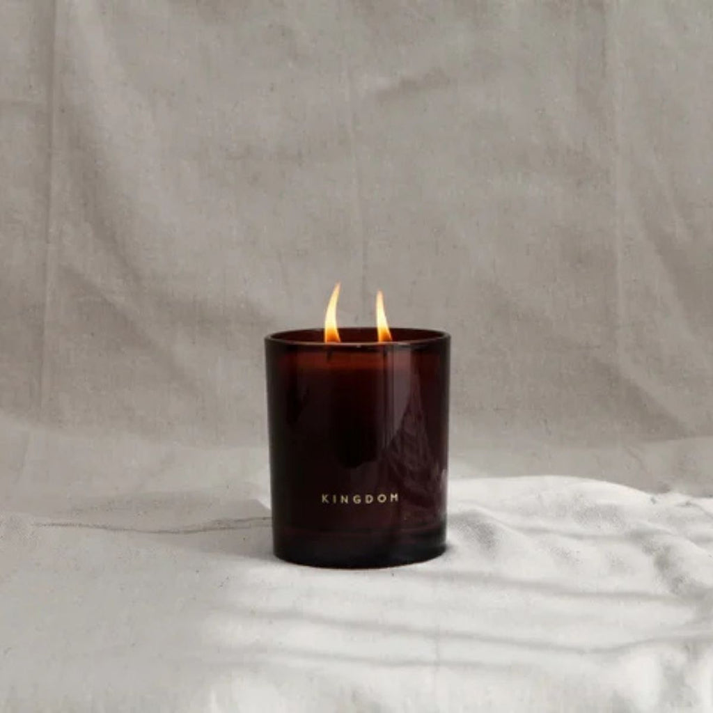 Candle in a dark glass container labeled 'KINGDOM' on a light fabric background