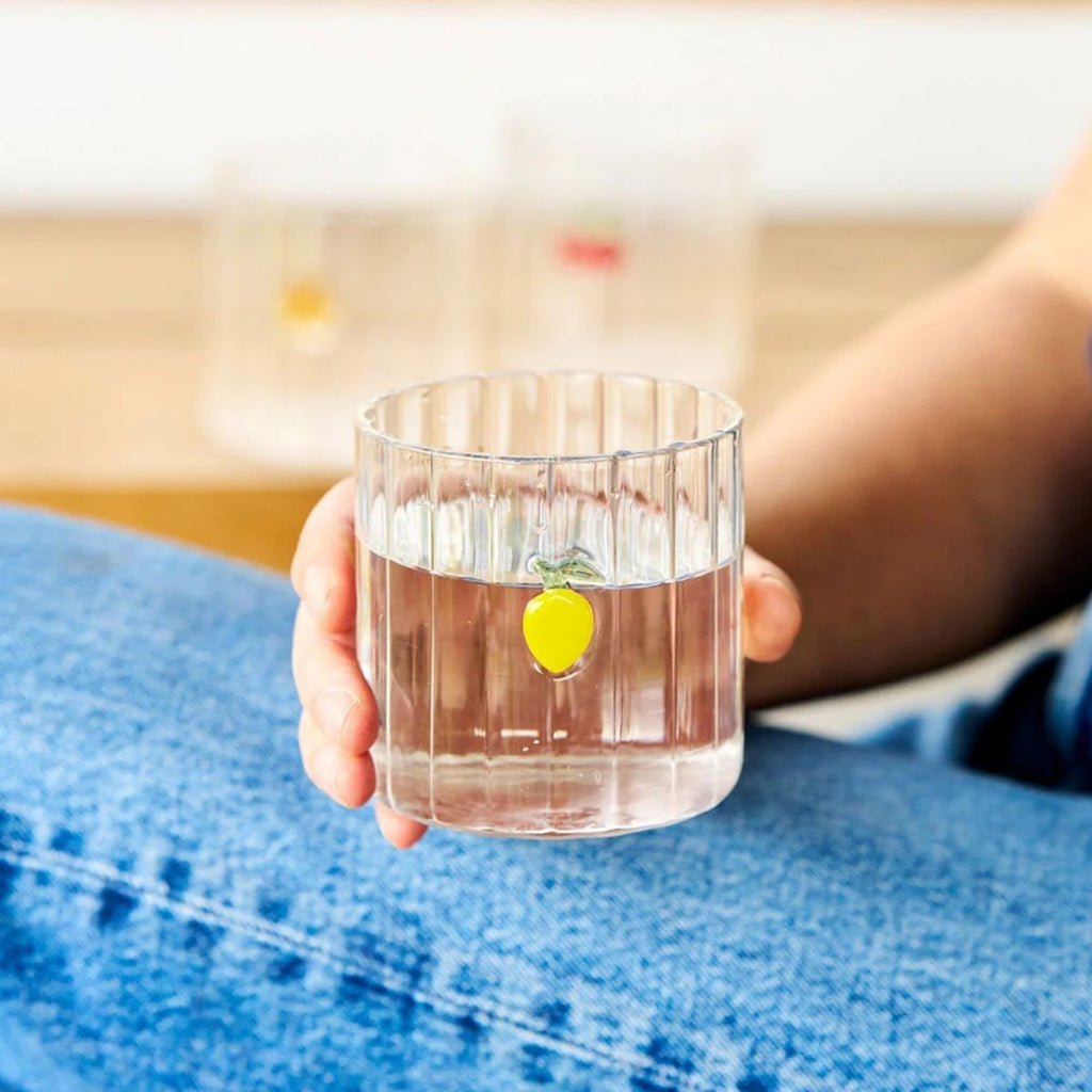 Hand holding a glass of water with a lemon slice, sitting on a blue surface.