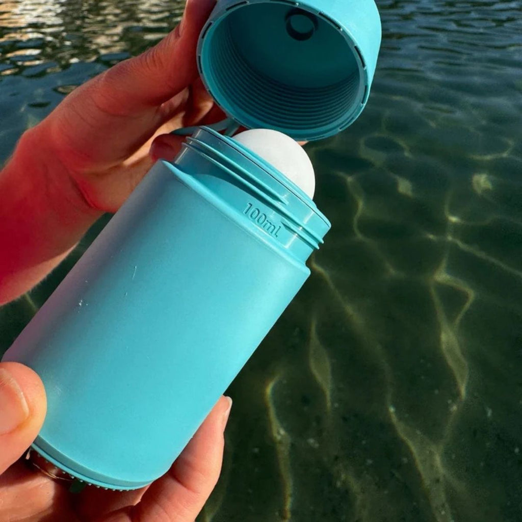 Hand holding a blue silicone sunscreen with a cap over clear water