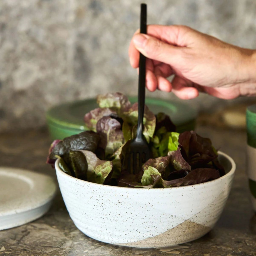 Person holding a black fork over a bowl of leafy greens on a stone surface.