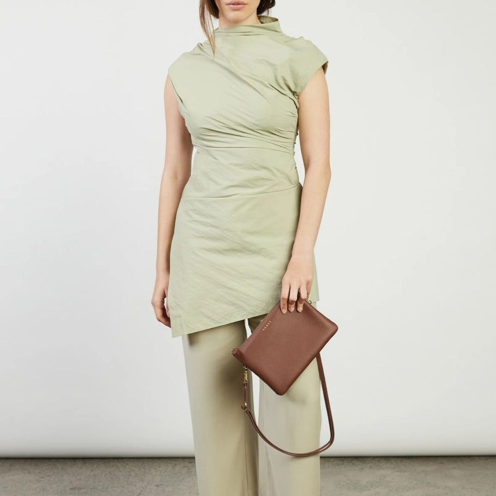 Woman wearing a light green sleeveless top and beige pants, holding a brown handbag against a white background.