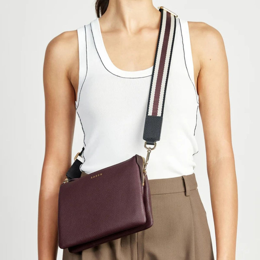 Person wearing a white tank top and brown pants with a maroon handbag on a plain background