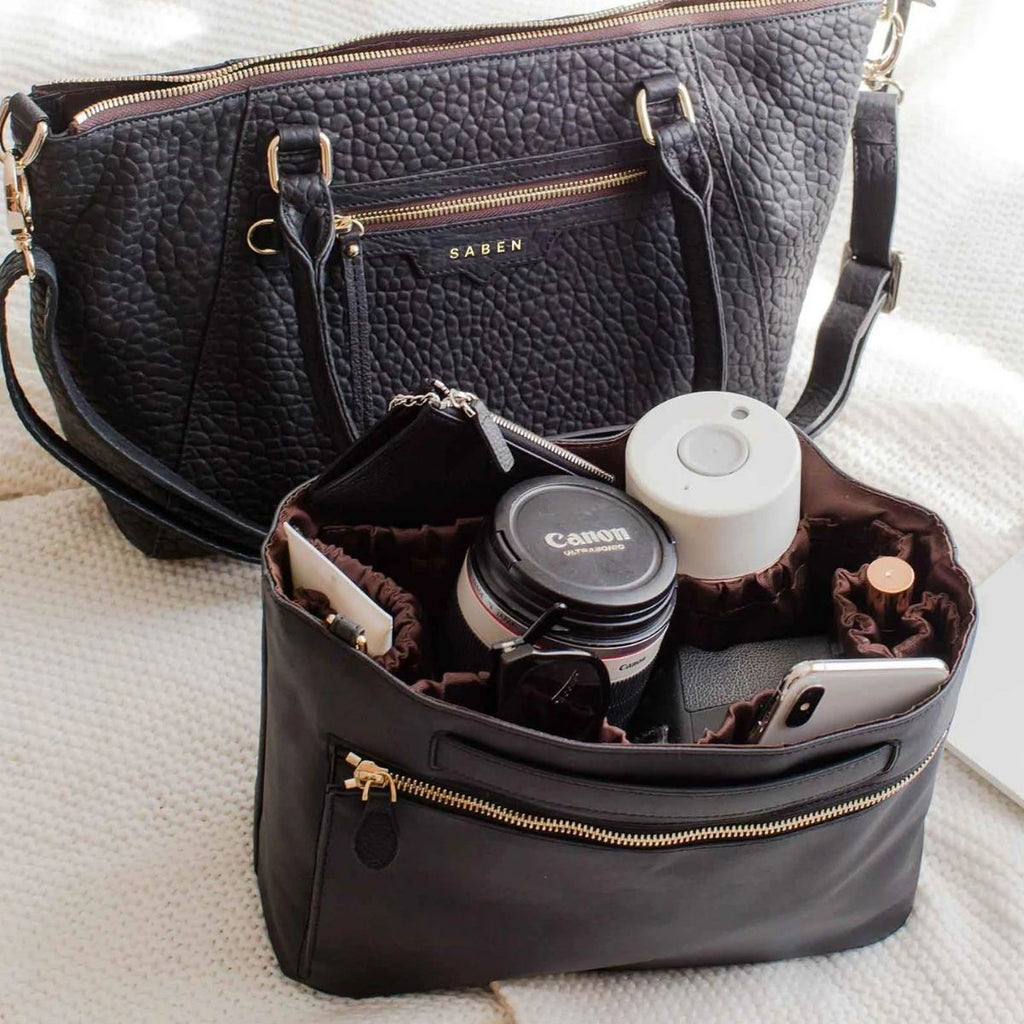 Black handbag with gold zippers containing a camera, bottle, and phone on a light surface.