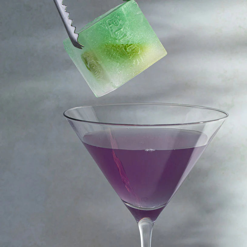 Green ice cube being placed into a purple cocktail in a martini glass.