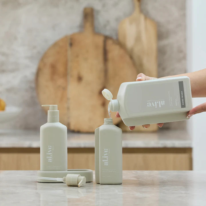 Person pouring liquid from a white bottle labeled 'alive' into a beige bottle on a kitchen counter.