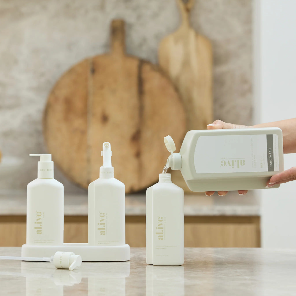 Person pouring liquid from a bottle into other bottles on a kitchen counter with wooden cutting boards in the background.