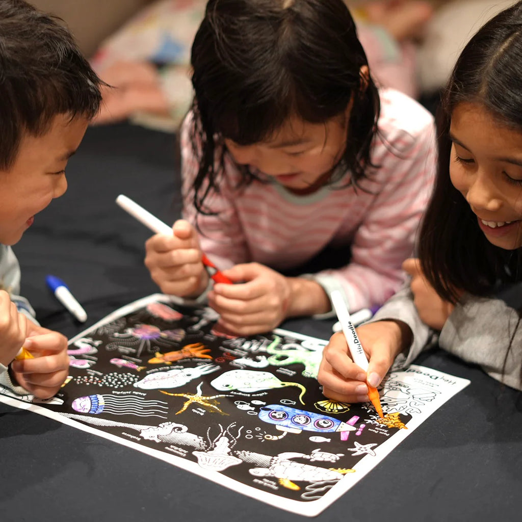 Three children coloring on a black paper with markers.