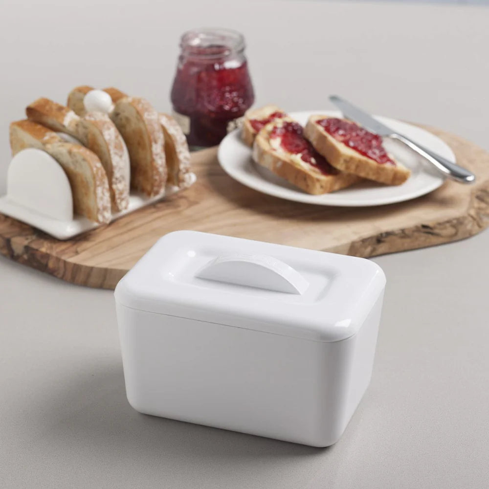 White ceramic butter dish with bread and jam on a wooden board in the background