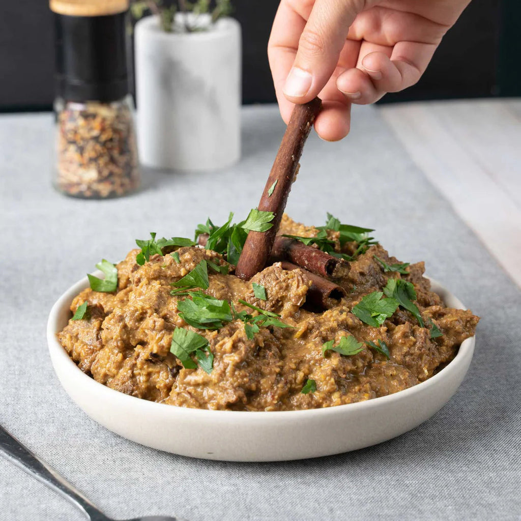 Hand using a wooden spoon to serve a dish of food garnished with herbs, placed on a gray surface with a blurred background.