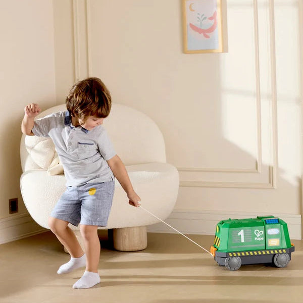 Child pulling a toy garbage truck in a room with a white chair and framed picture on the wall.