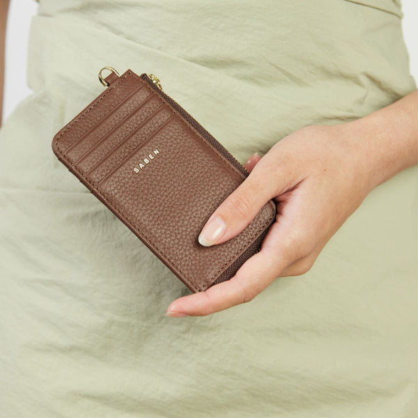 Brown wallet with 'SABEN' branding held by a hand against a light green background