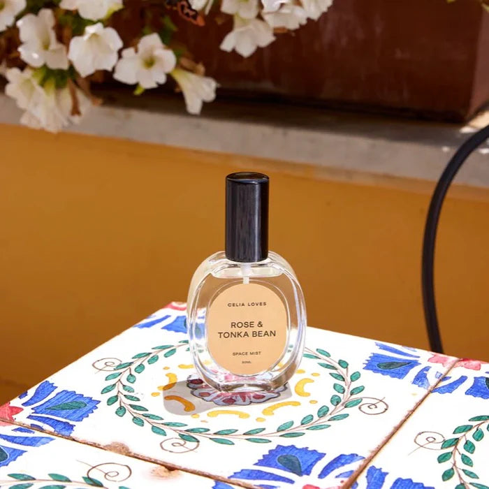 Room Spray bottle labeled 'Rose & Tonka Bean' on a decorative table with flowers in the background