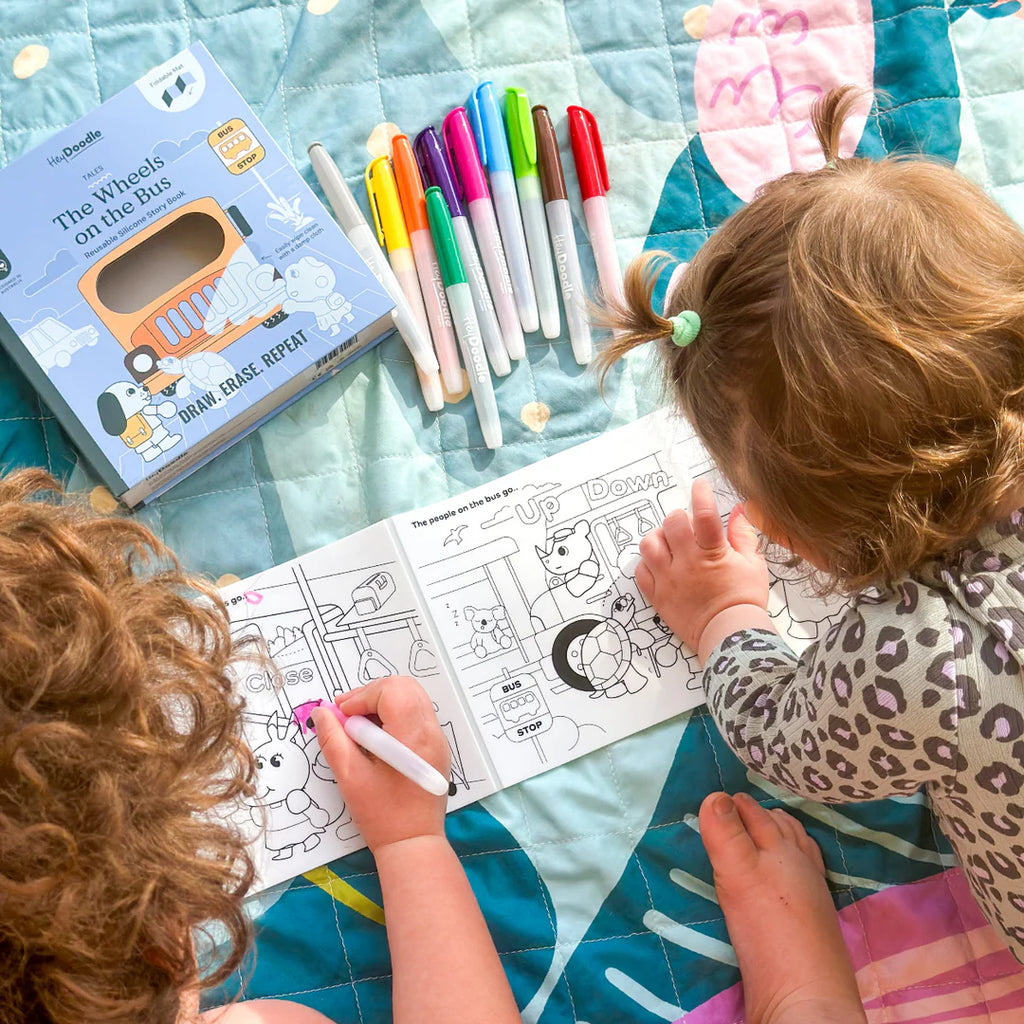 Two children coloring with markers on a blanket, with a book titled 'The Wheels on the Bus' nearby.