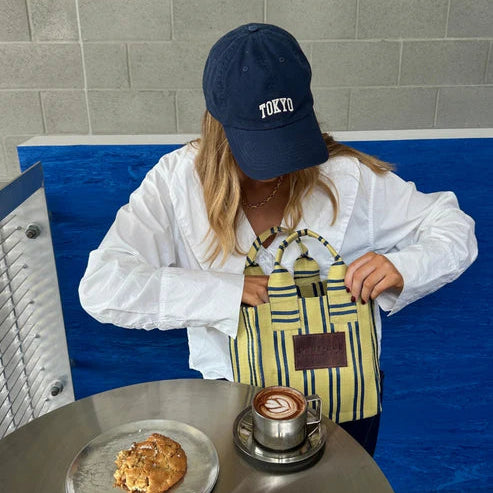 Person holding a striped bag with a cup of coffee and cookie on a table, wearing a 'Tokyo' cap.