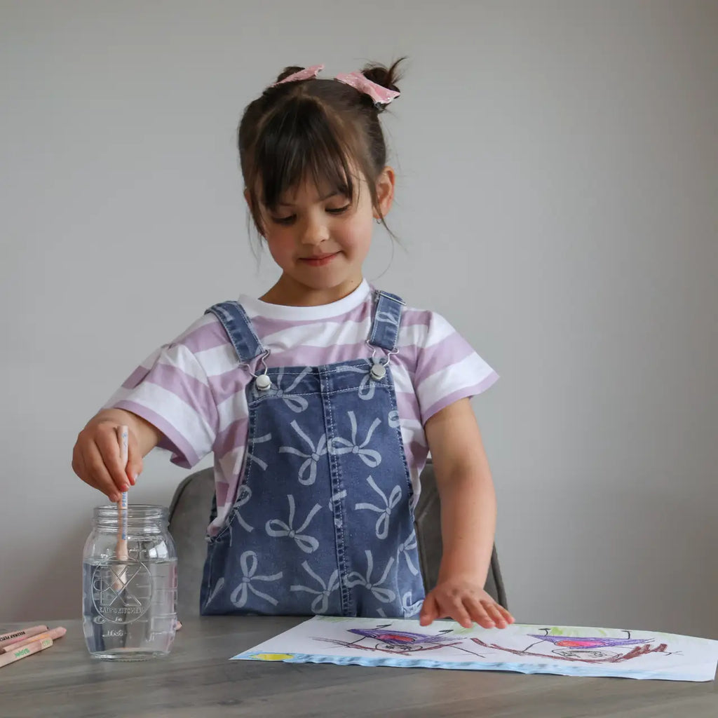 Child in a striped shirt and denim overalls sitting at a table with a coloring book and crayons.
