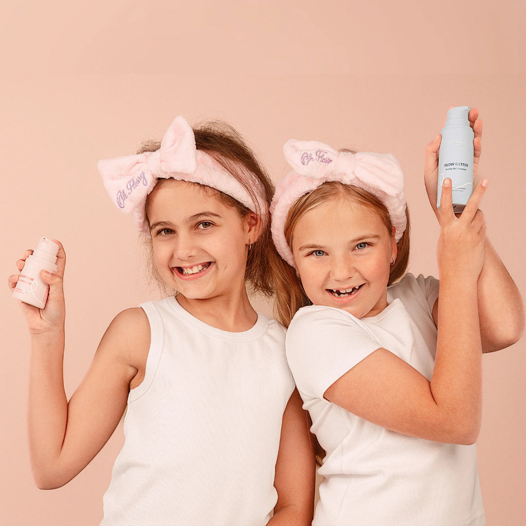 Two children wearing pink headbands with bows, holding skincare products against a beige background.