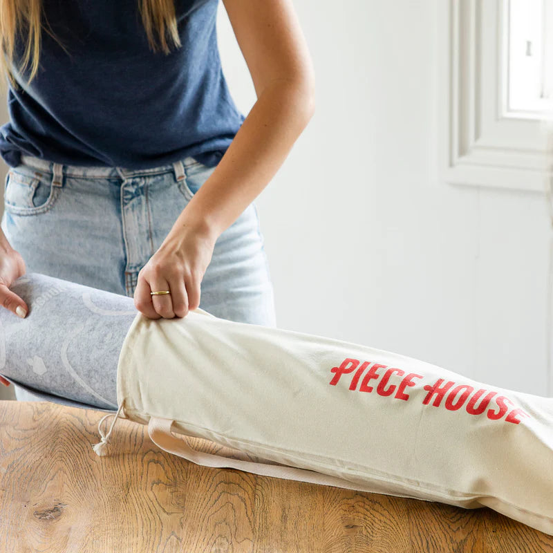 Person unrolling a piece of fabric from a 'PIECE HOUSE' branded bag on a wooden floor.