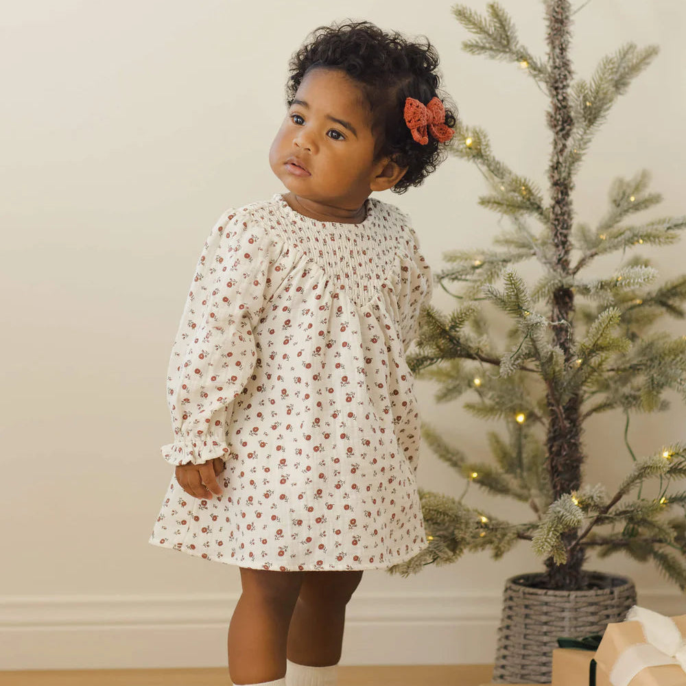 Young child in a floral dress standing next to a decorated Christmas tree with presents.