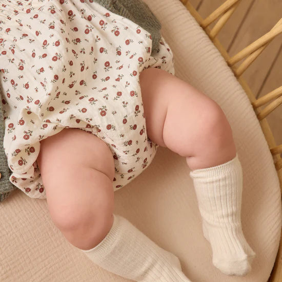 Baby wearing white socks and a floral outfit sitting on a textured surface.