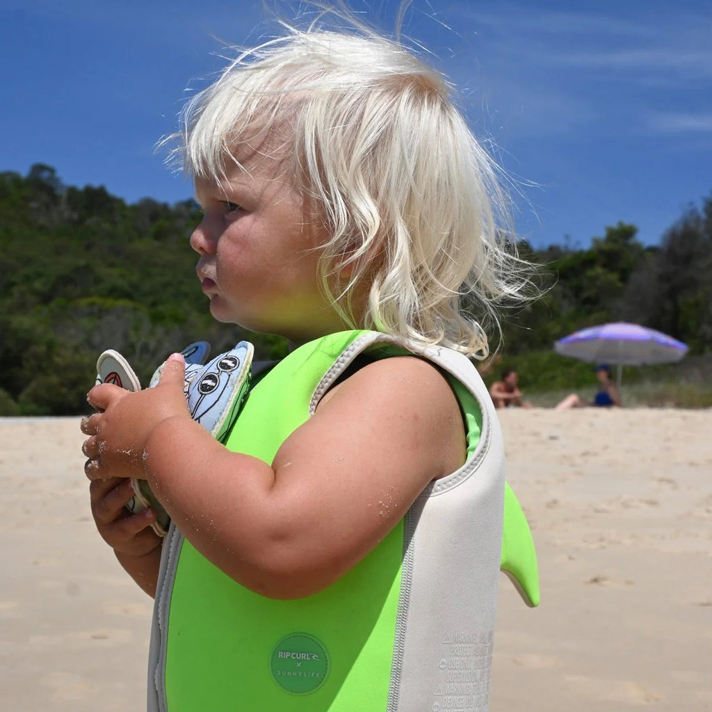 Child in a green life jacket holding flipper shoes on a sandy beach with trees and an umbrella in the background.