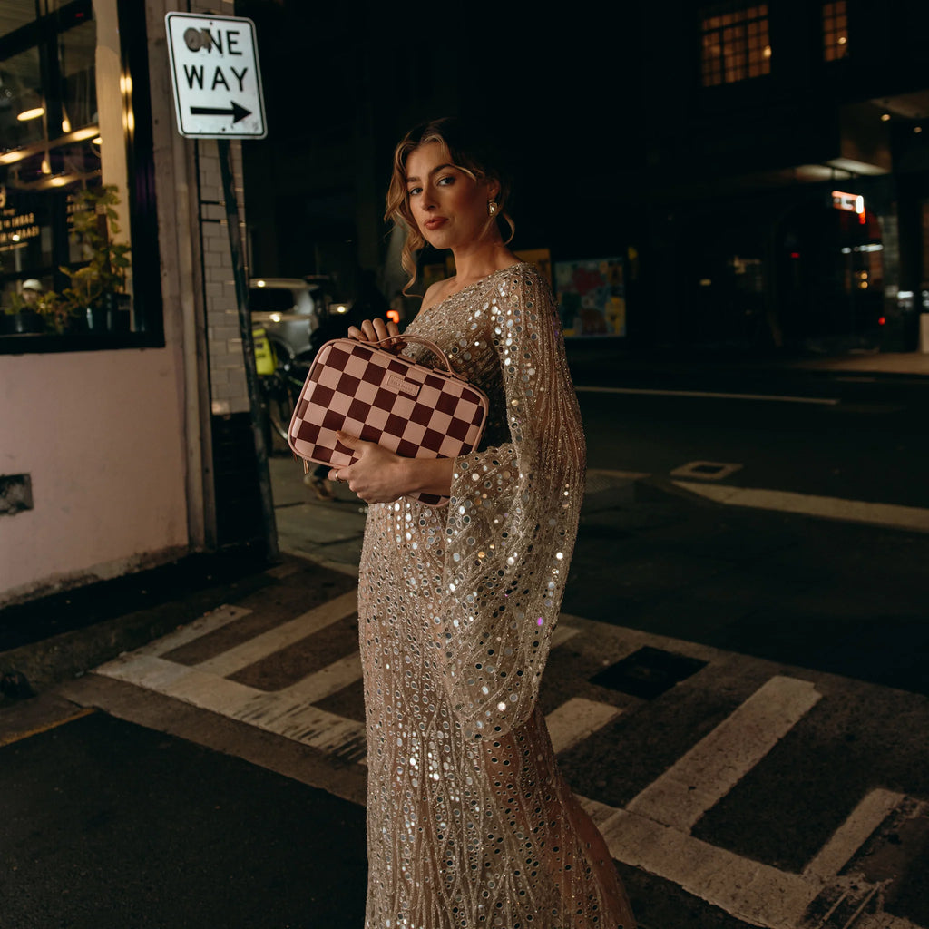 Woman in a sparkling dress holding a checkered bag on a city street at night.
