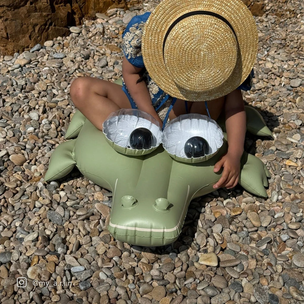 Child sitting on a green inflatable crocodile toy on pebbles
