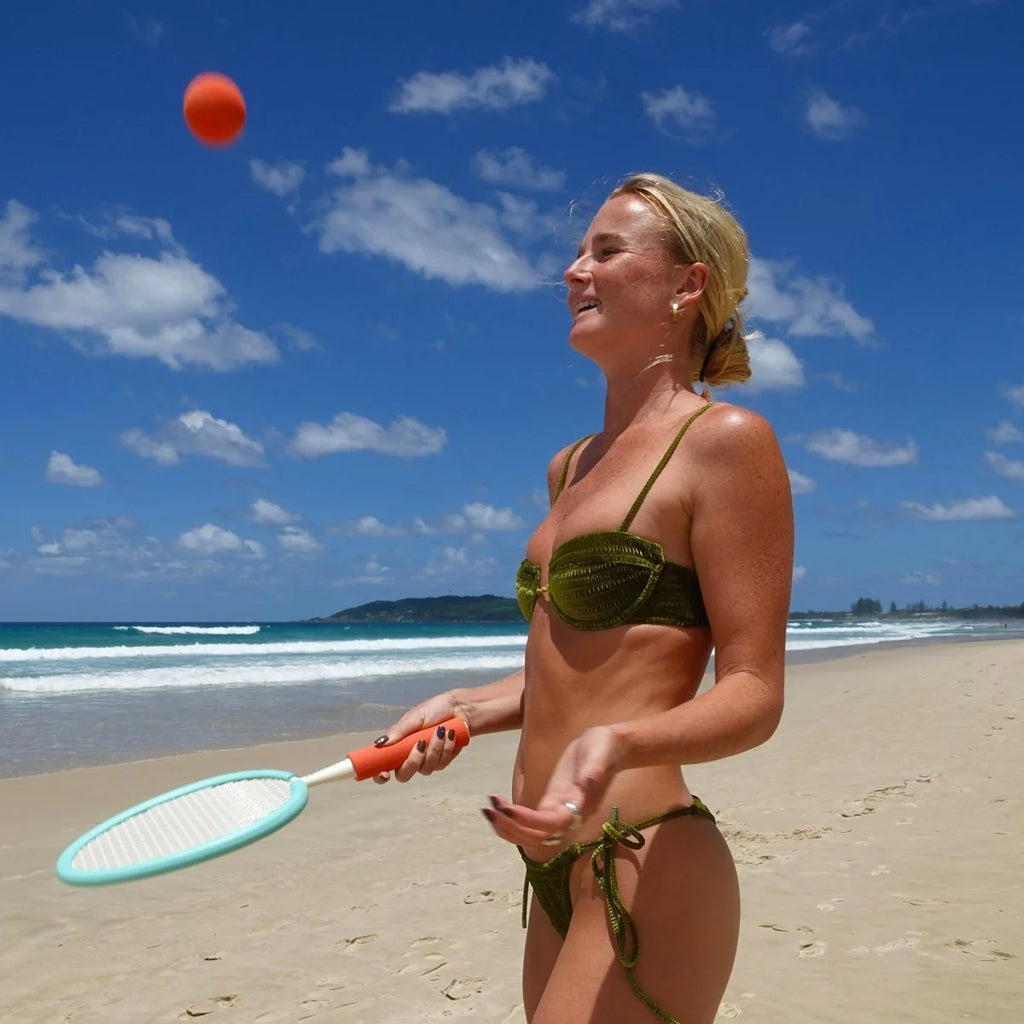Woman in green bikini playing beach tennis with a red ball on a sunny beach.
