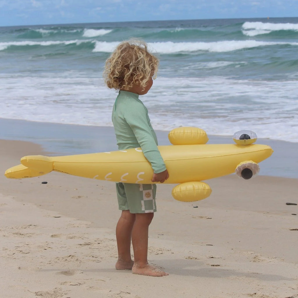 Child holding a yellow inflatable shark toy on a beach with ocean waves in the background