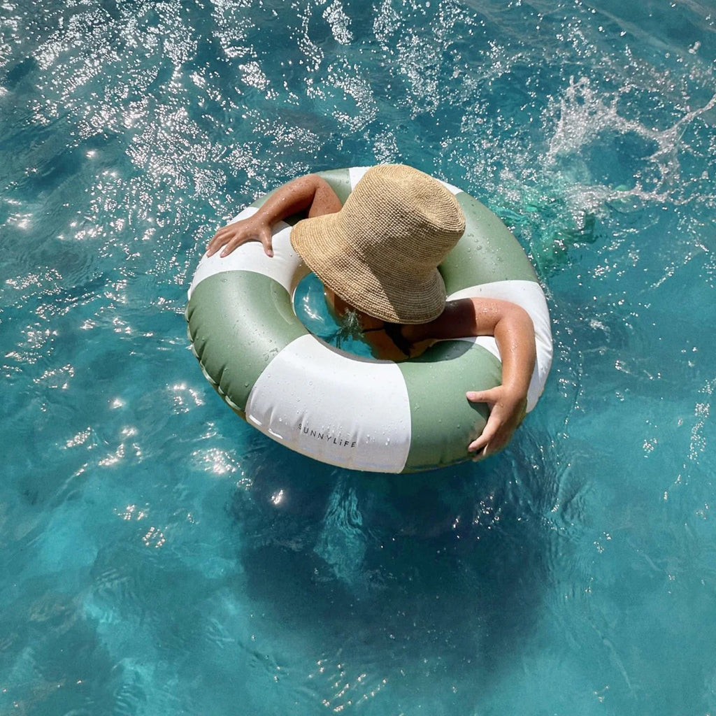 Person wearing a straw hat and floating on a green and white inflatable ring in clear blue water.