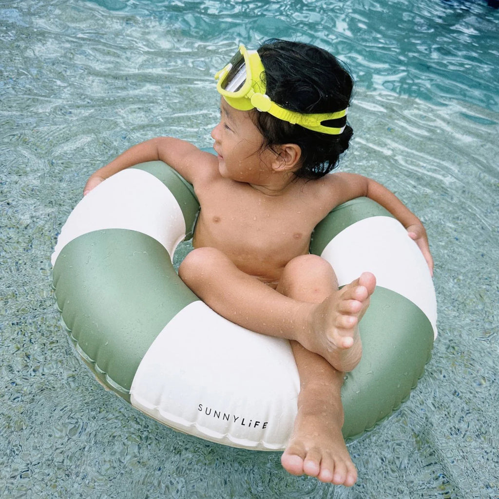 Child sitting on a green and white inflatable ring in a pool, wearing goggles.
