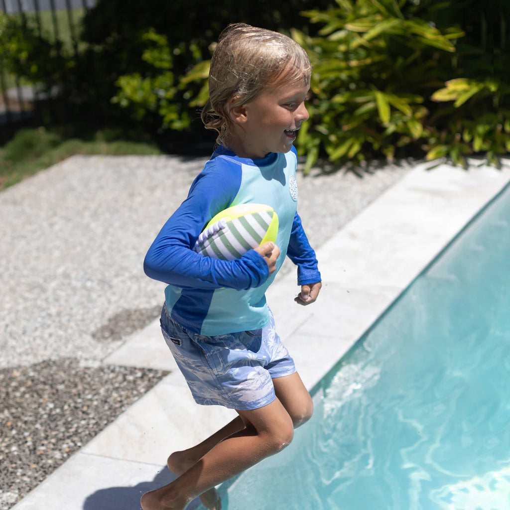 Child in a blue and light blue swimsuit holding a striped ball by a pool.