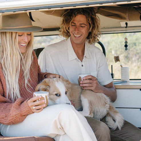 Two people with a dog inside a vehicle, holding mugs.
