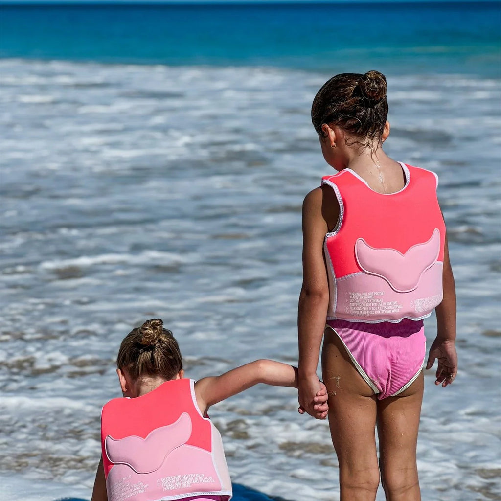 Two children in pink life vests standing on a beach with ocean waves in the background