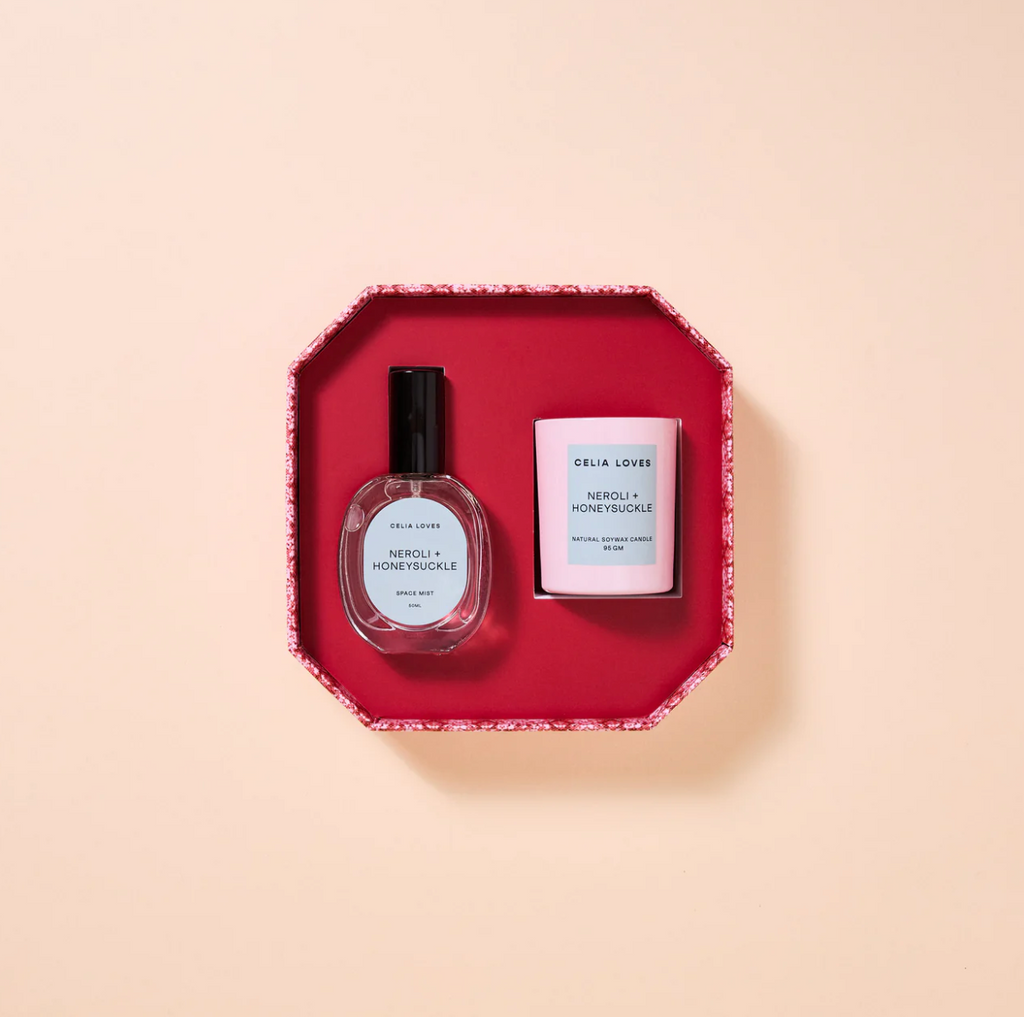 Two beauty products, a room spray bottle and a candle, on a pink box with a beige background.