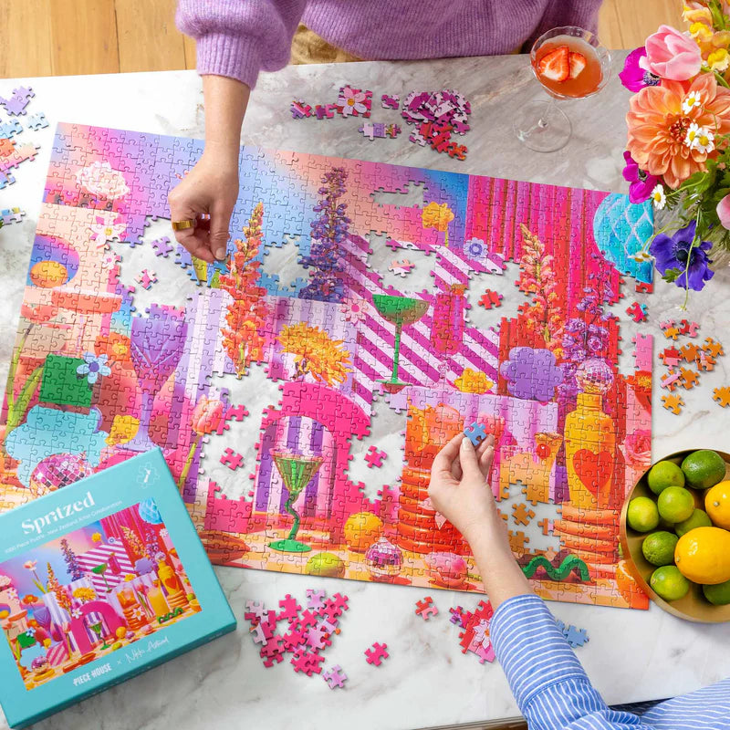 Two people working on a colorful puzzle with a 'Spooked' box, flowers, and fruit on a table.