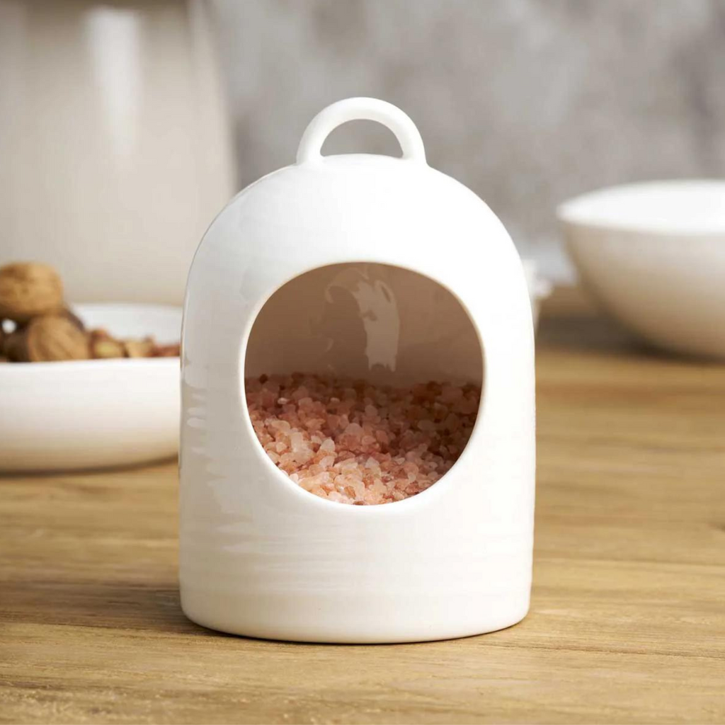 A white ceramic salt pig filled with Himalayan salt on a wooden table with two white ceramic bowls in the background 