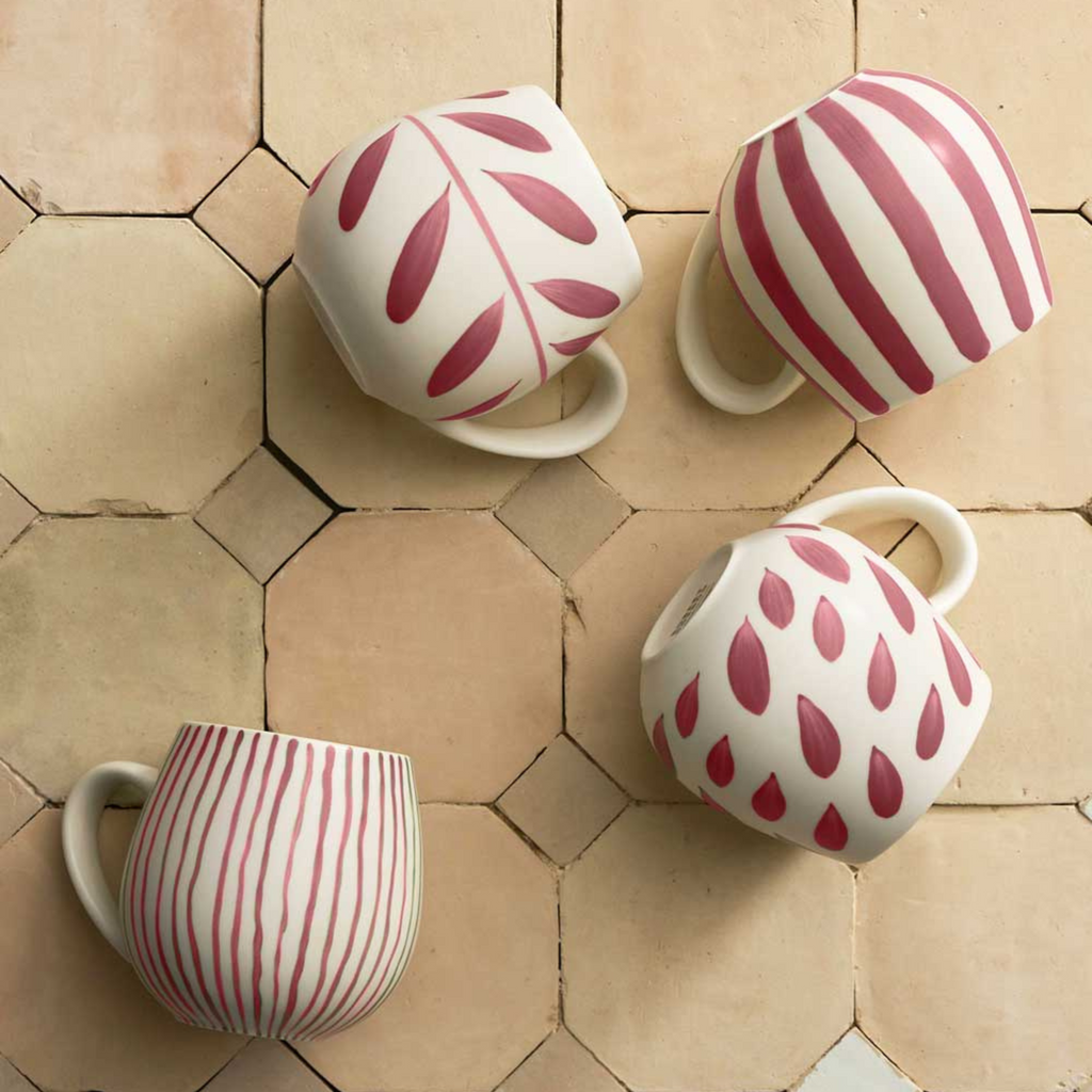 A set of four rounded shape mugs with various painted patterns in pink paint with a handle rested on a beige tile background