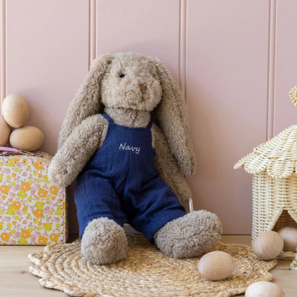 A plush toy rabbit with beige fur and navy blue overalls, against a pink wall background and eggs scattered around