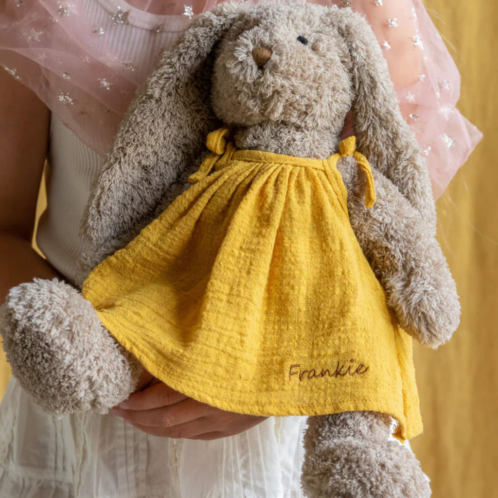 A plush toy rabbit with a beige body and long ears, wearing a mustard yellow dress with the text Frankie' embroidered on it.