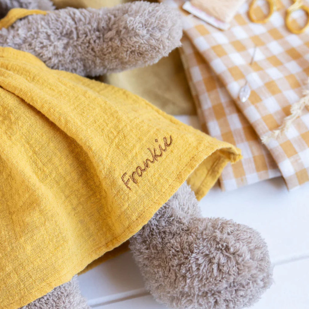 close up of a  plush toy rabbit with a beige body and long ears, wearing a mustard yellow dress with the text Frankie' embroidered on it.