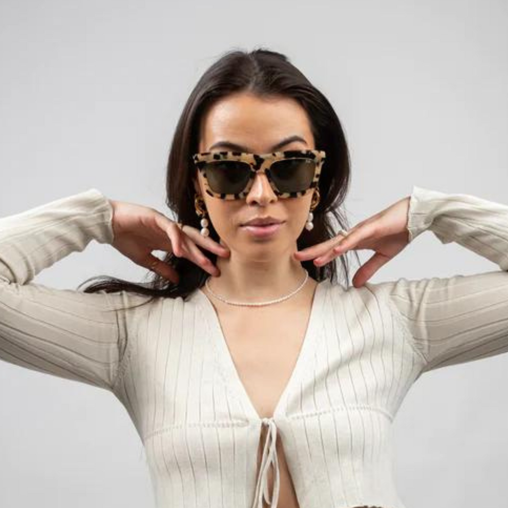 A woman wearing sunglasses with a solid tortoise frame and dark lens framing her face with her hands in a white top