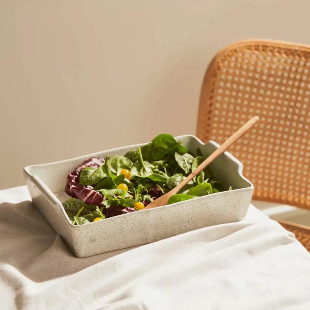 A  rectangle white and black speckled ceramic serving dish holding a salad and wooden spoon on a table with a table cloth with a rattan chair pushed under it.