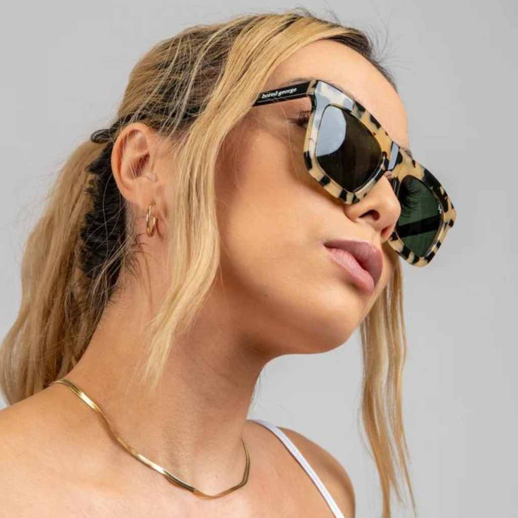 A woman wearing sunglasses with a solid light tort frame and dark lens and a gold chain looking to the side of the camera