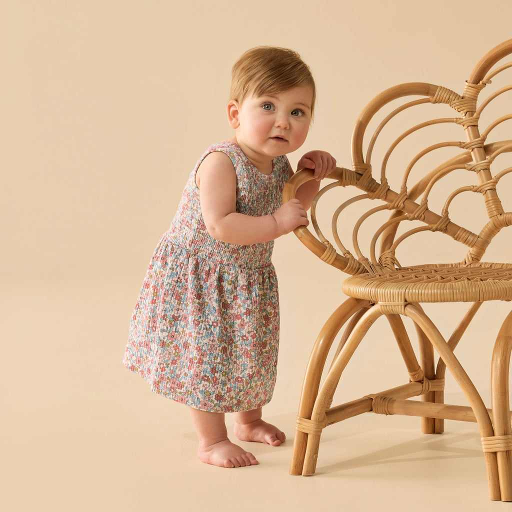 Baby in a floral dress standing next to a wicker chair on a beige background