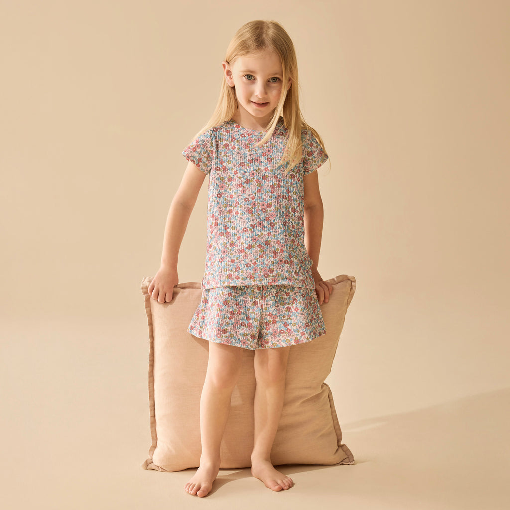 Young girl wearing a floral pajama set standing next to a beige pillow on a beige background