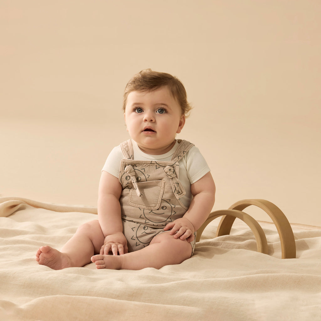 Baby sitting on a beige blanket wearing a patterned romper with a wooden toy.
