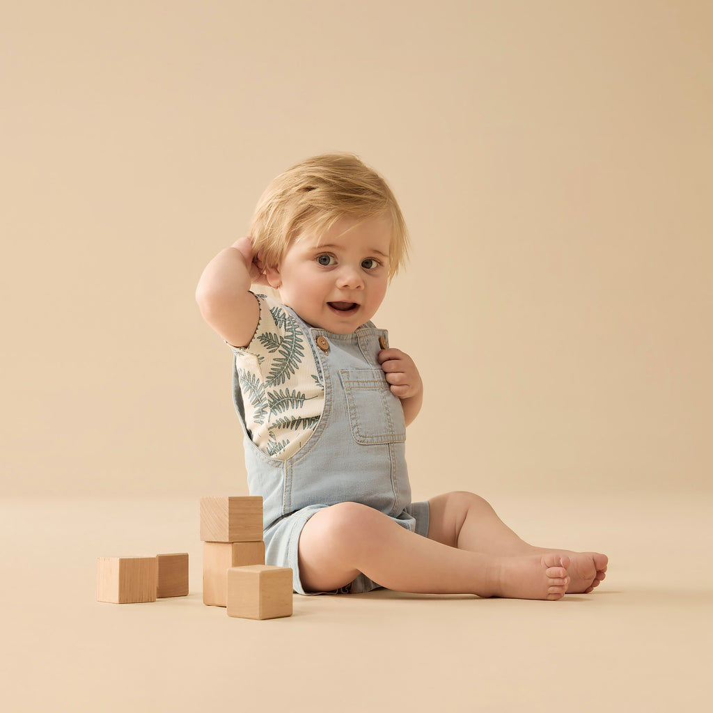 Baby sitting on a beige surface with wooden blocks and wearing a bib and overalls.
