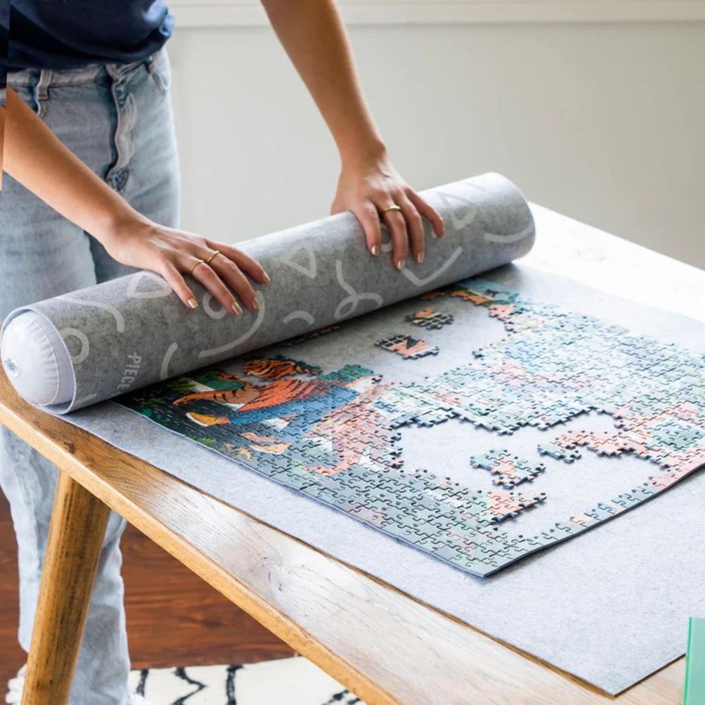 Person unrolling a puzzle mat with a partially completed puzzle on a wooden table.